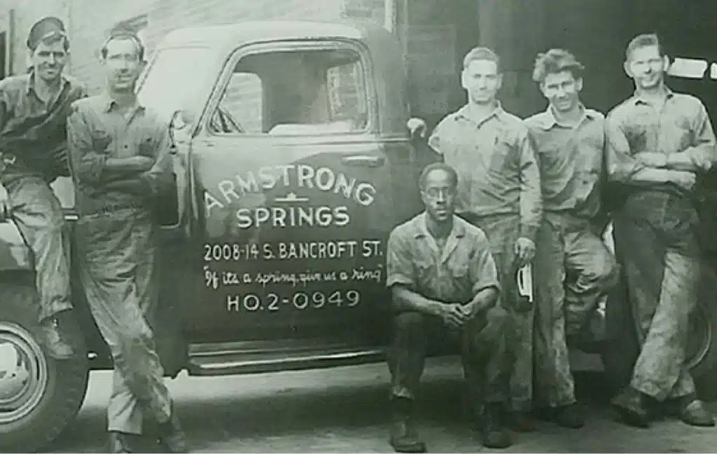 A group of six men in work uniforms pose beside a truck labeled "Armstrong Springs," showcasing their pride and professionalism like a homepage banner. The vehicle is prominently marked with a phone number and address, symbolizing their commitment to service.
